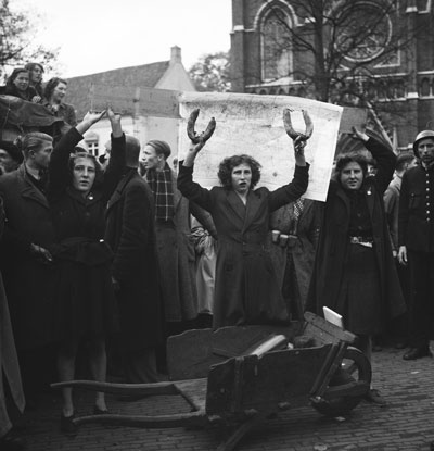 "Moffenmeiden" op De Heuvel te Tilburg, november 1944. Twee vrouwen houden een plank met een kaart omhoog. Daarvoor een andere vrouw die twee worsten omhoog houdt. (Bron: Nationaal Archief, coll. Anefo, nr. 934-9738. CCO 1.0 Universeel / Publiek Domein)
